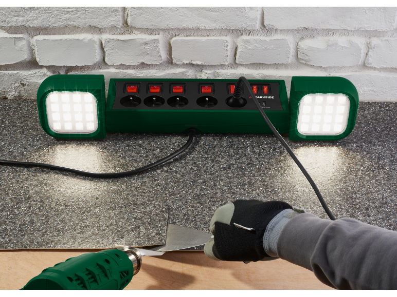Stekkerdoos met geïntegreerde LED-lampen, gebruikt met een heteluchtpistool en schraper op een vloer.