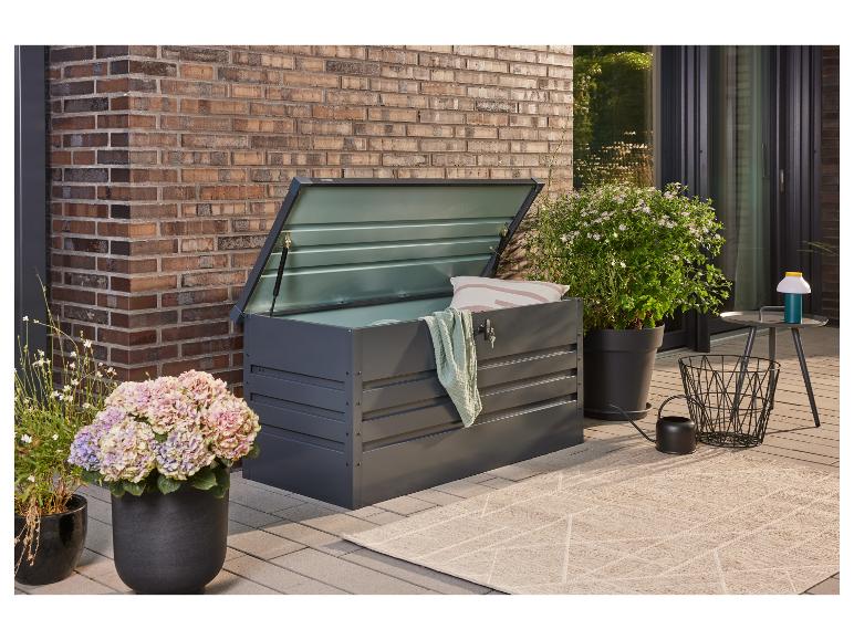 Donkergrijze tuinopbergbox met open deksel, kussen en deken op een terras.