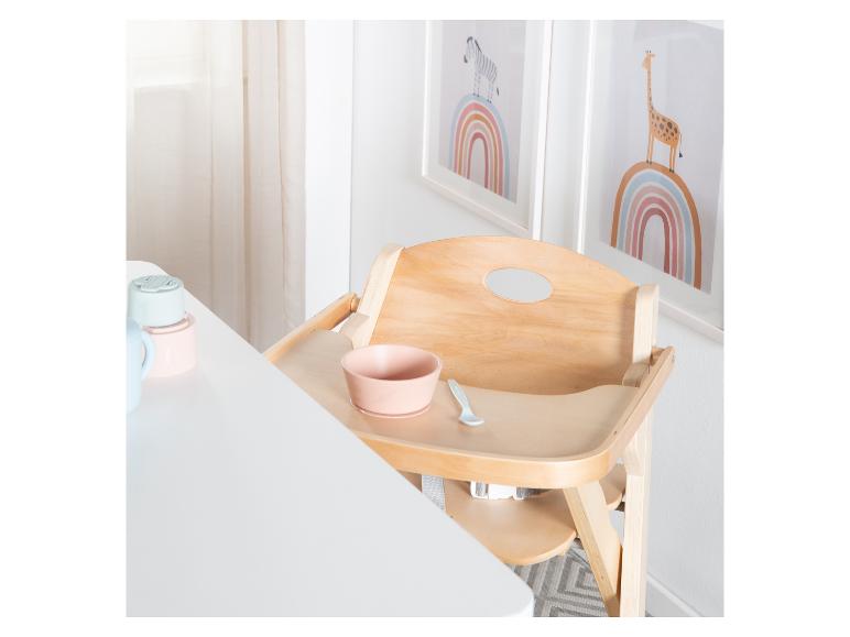 Houten kinderstoel met een roze kom en lepel, naast een witte tafel met babyservies.