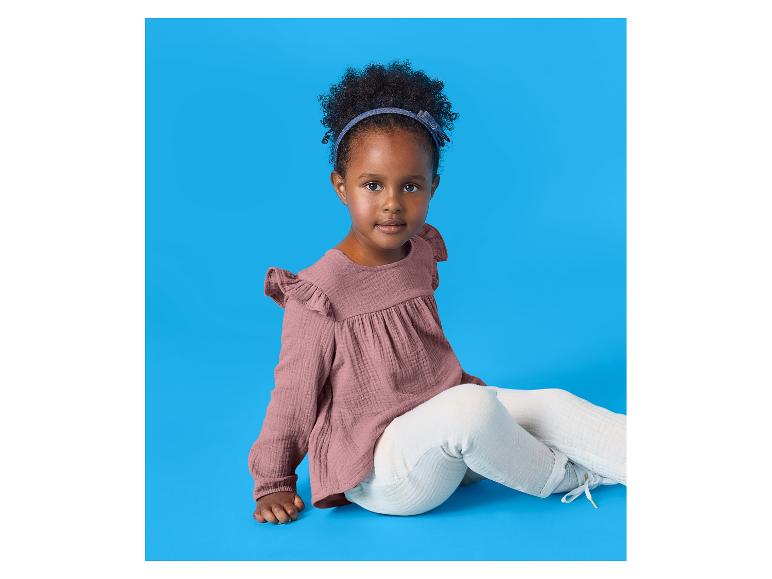 Meisje met krullend haar, roze blouse en witte broek, zittend op een blauwe achtergrond.
