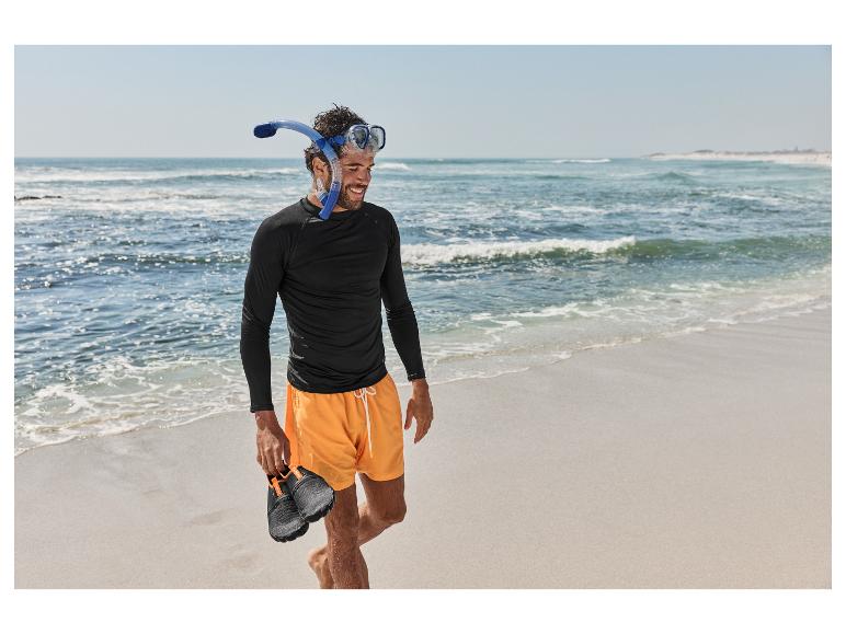 Man in zwart shirt, oranje korte broek, snorkelmasker en waterschoenen op het strand.
