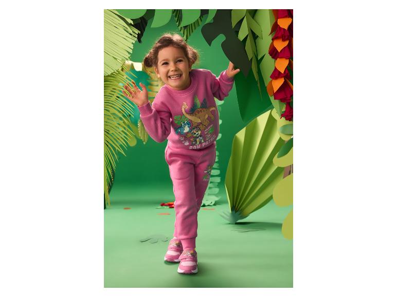 Meisje in roze trainingspak met dinosaurusprint en sneakers, lachend in een papieren jungle-setting.