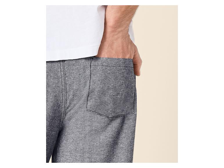 Man in een wit T-shirt en grijze korte broek, met zijn hand in zijn zak.