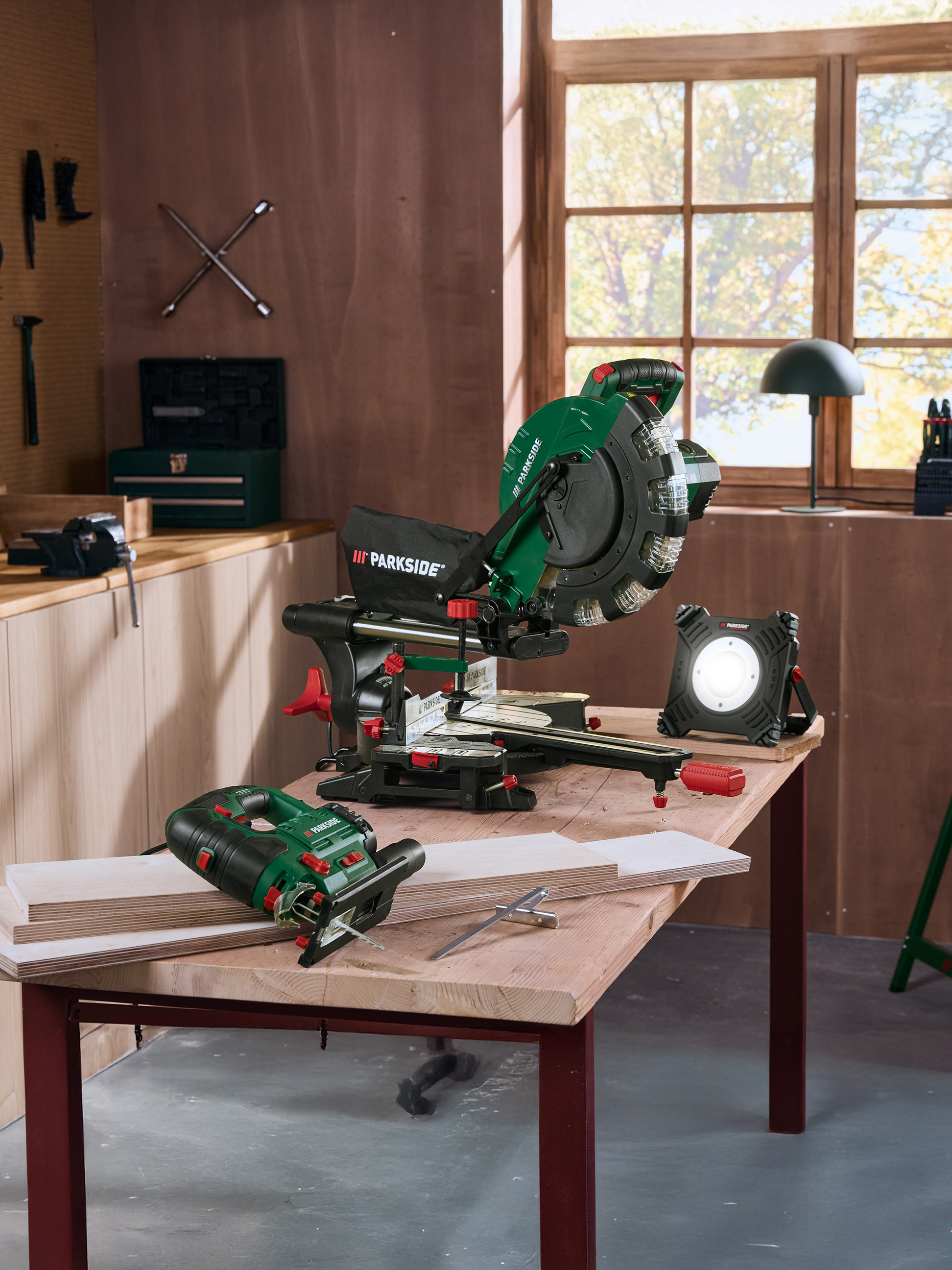 Parkside gereedschap: afkortzaag, decoupeerzaag en bouwlamp op een houten tafel in een werkplaats.