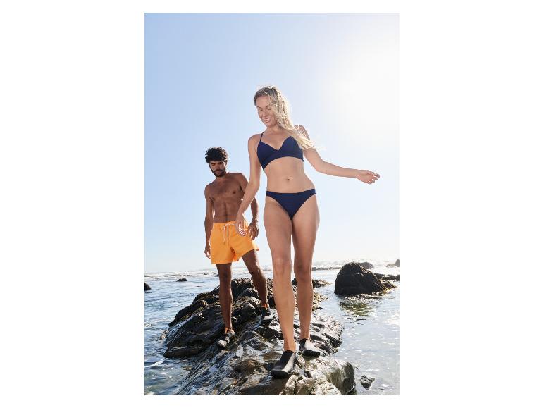 Vrouw in donkerblauwe bikini en man in oranje zwemshort op rotsachtig strand.