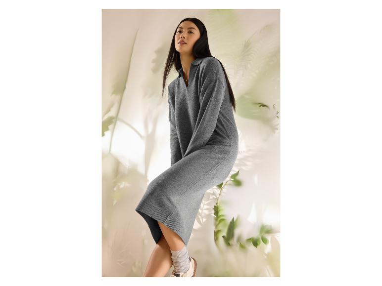 Vrouw in een grijze gebreide jurk en witte sneakers, zittend tegen een lichte achtergrond met planten.