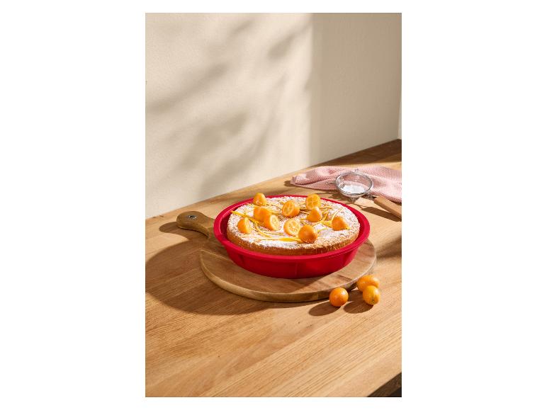 Ronde cake versierd met kumquats en citroenschil in een rode bakvorm op een houten plank.