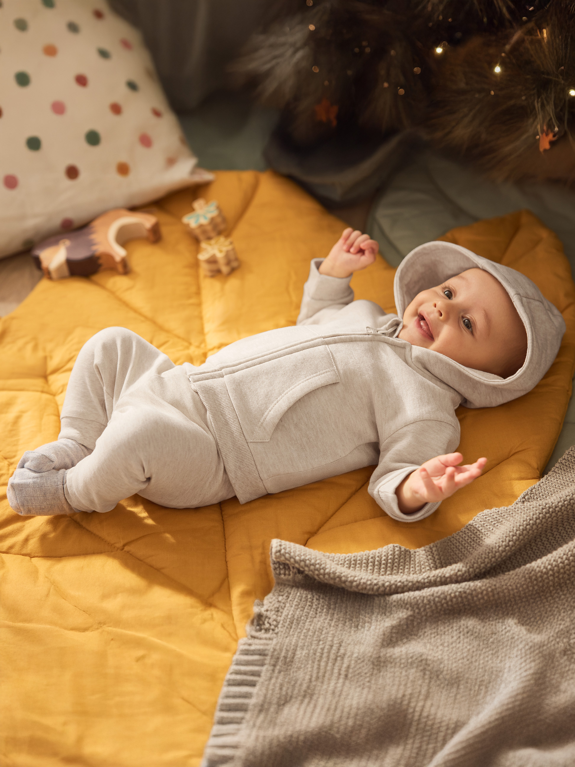 Blije baby in grijs pak op geel matras met speelgoed en kerstverlichting.