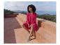 Vrouw in fuchsia jurk en comfortabele schoenen zittend op een terras met zeezicht.