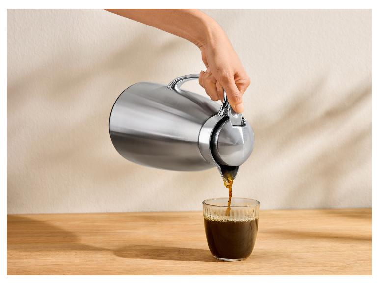 Hand schenkt koffie uit zilveren thermoskan in glazen beker op houten tafel.