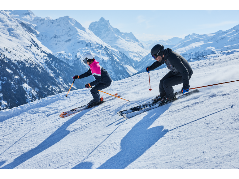 Twee skiërs in skikleding en helmen skiën van een besneeuwde berghelling.