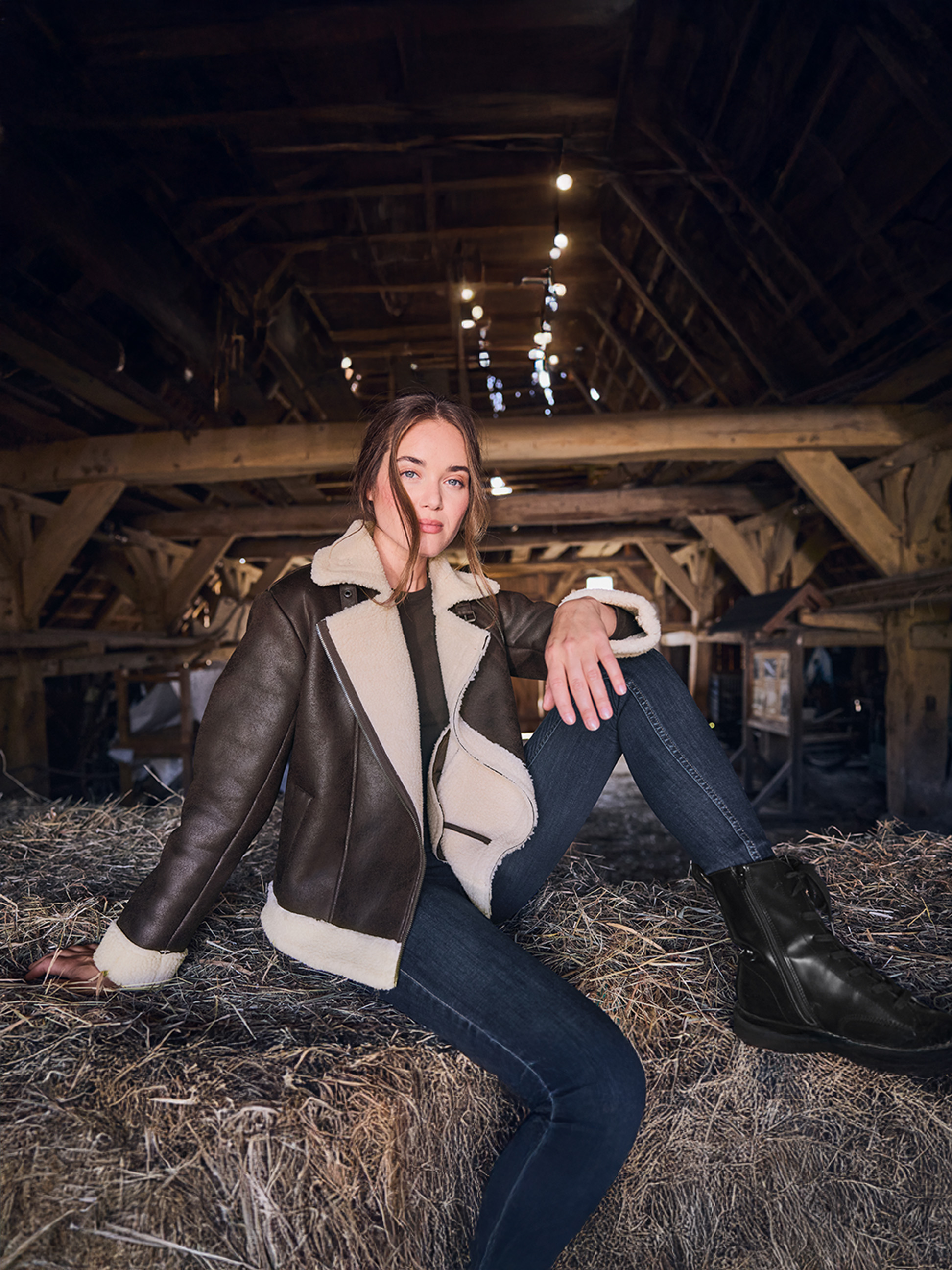 Vrouw in een bruine shearling jas en jeans, zittend op hooibalen in een schuur.