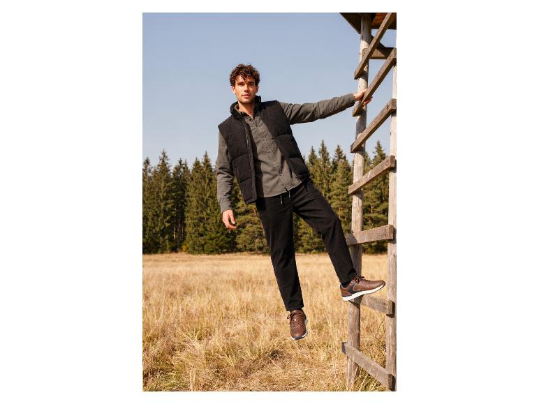 Man met spijkerbroek, vest en bruine sneakers op een houten ladder in de natuur.