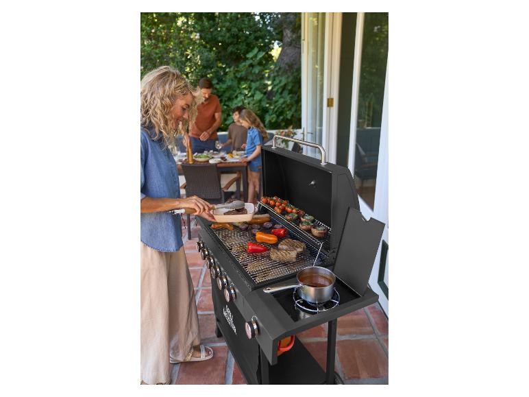 Vrouw grilt vlees en groenten op een gasbarbecue, met familie aan tafel op het terras op de achtergrond.