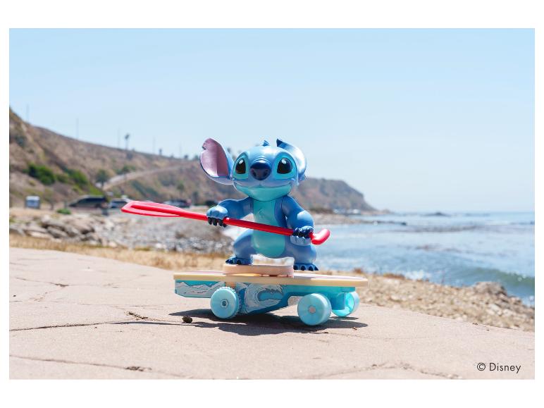 Stitch-figuur op skateboard met peddel, op een pad bij het strand.