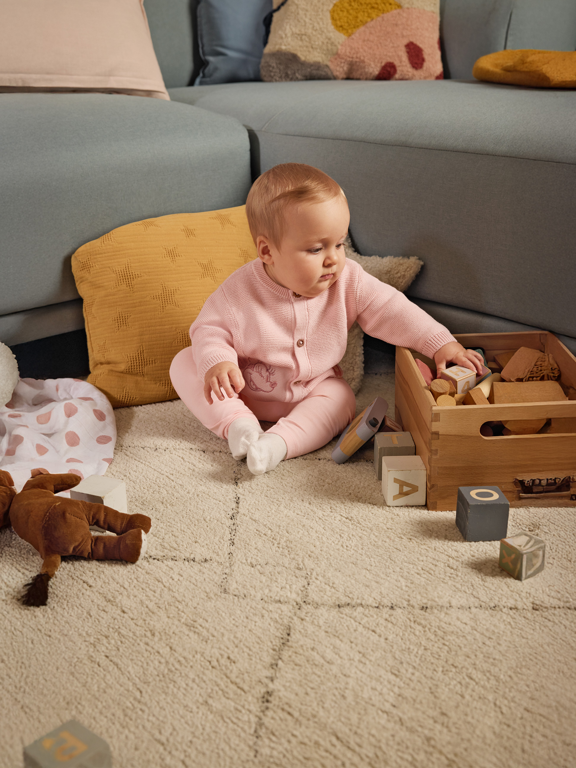 Baby in roze pak speelt met houten blokken op een tapijt naast een bank.