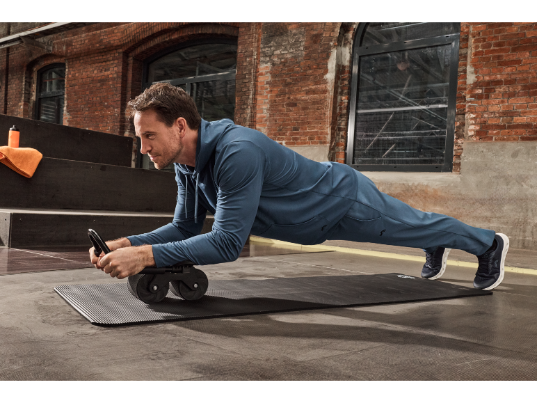 Een man doet een plankoefening op een fitnessmat met een buikspierwiel.