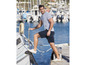 Man in gestreept shirt en korte broek staat op een boot in de haven.