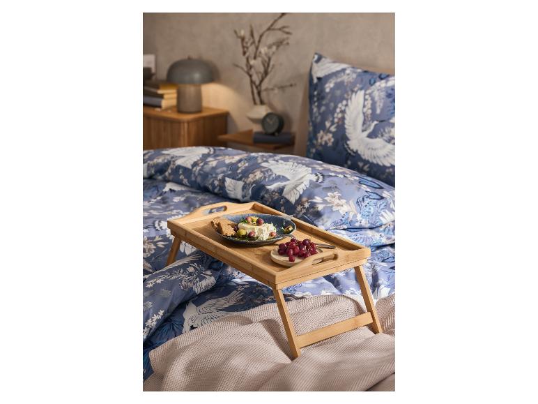 Een bamboe ontbijtblad met een bord eten en druiven op een bed met blauw-wit beddengoed met kraanvogels.