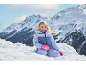 Meisje in skipak en handschoenen, lachend in de sneeuw met bergen op de achtergrond.