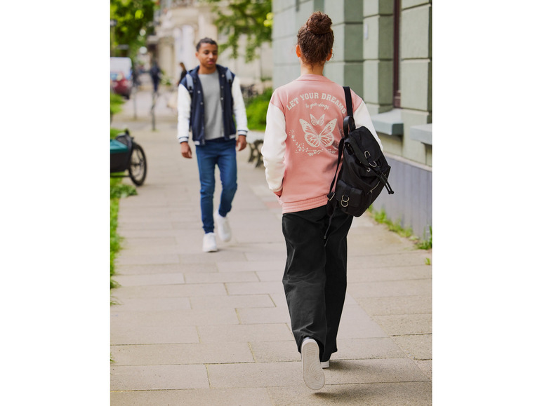 Een vrouw loopt op straat met een roze bomberjack en een zwarte rugzak.