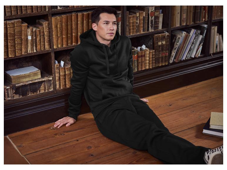 Man in zwarte hoodie en joggingbroek zit op de vloer naast een boekenplank.