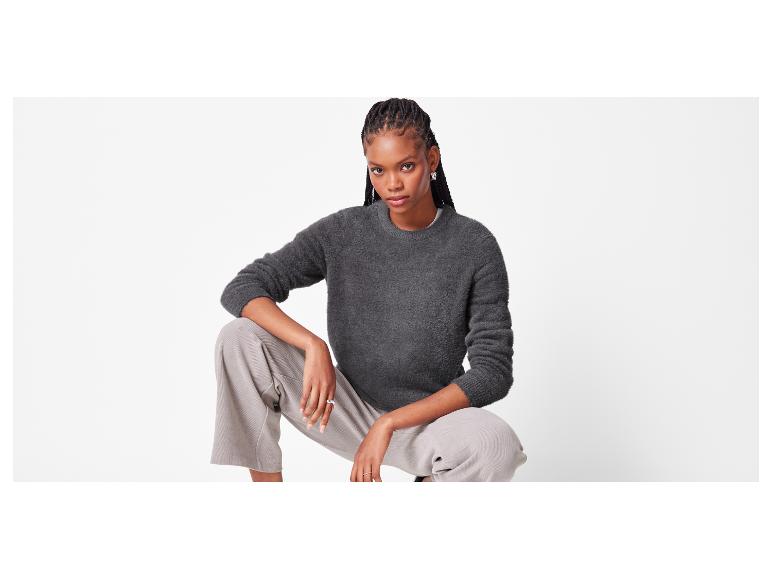 Vrouw in een donkergrijze pluizige trui en lichtgrijze broek gehurkt.