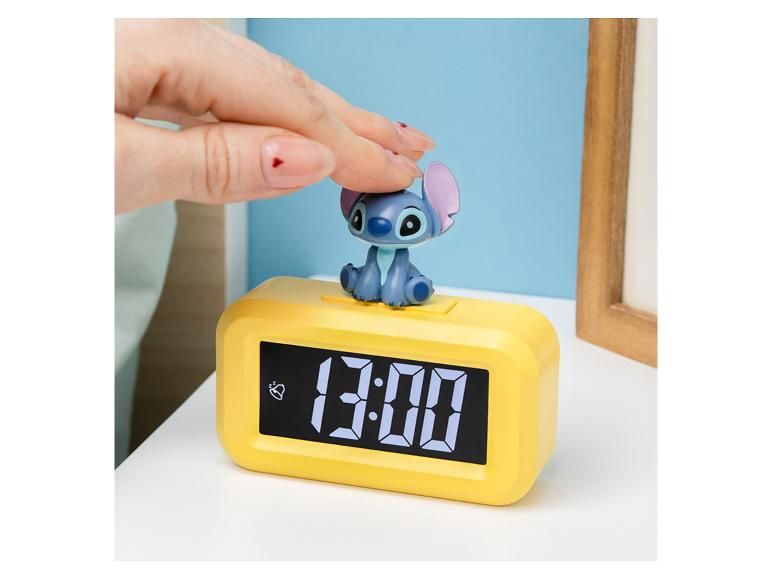Een hand drukt op een Stitch-beeldje bovenop een gele digitale wekker die '13:00' weergeeft.