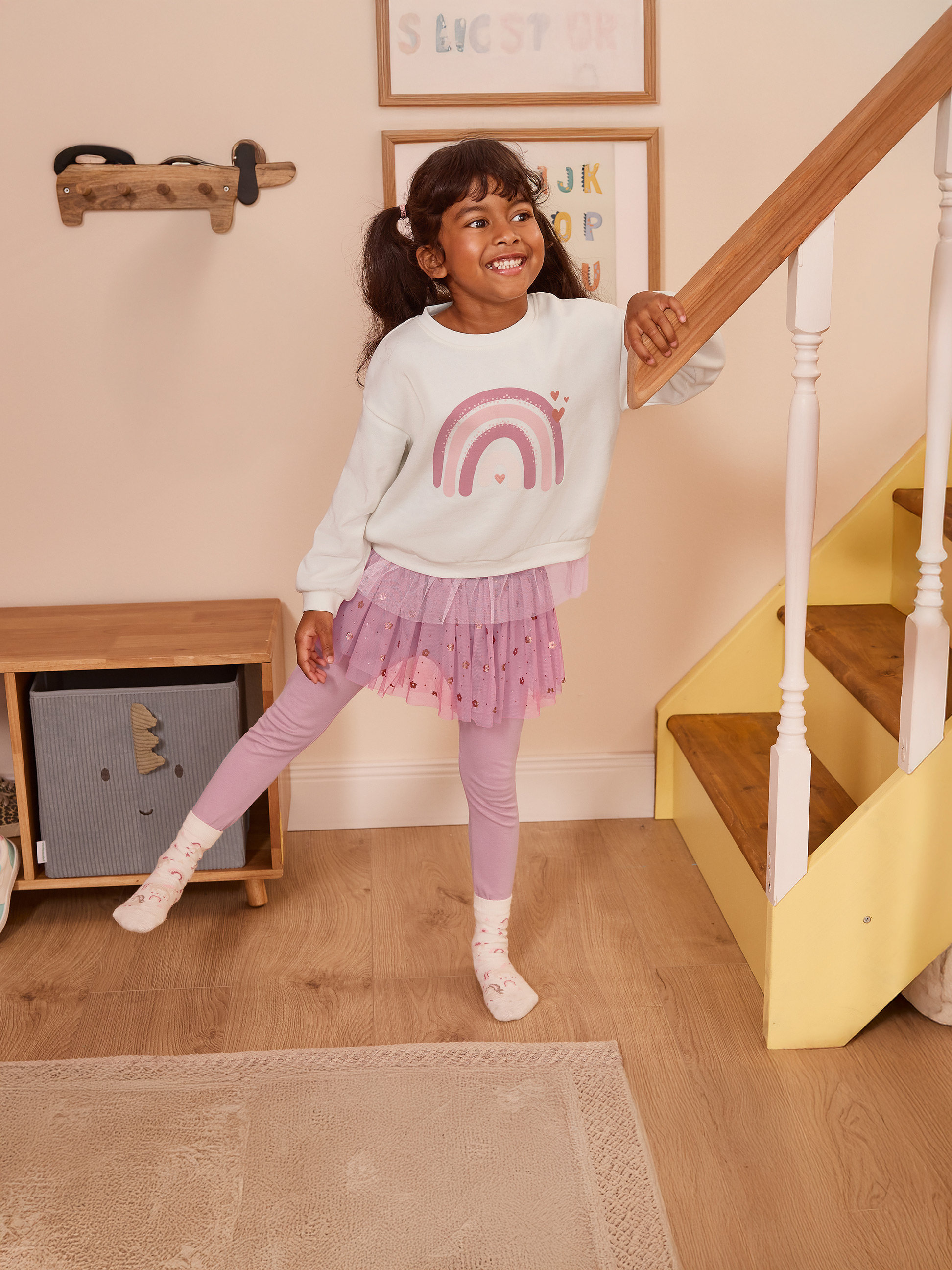 Meisje in roze tule rok en regenboog sweatshirt, leunend op een trapleuning.