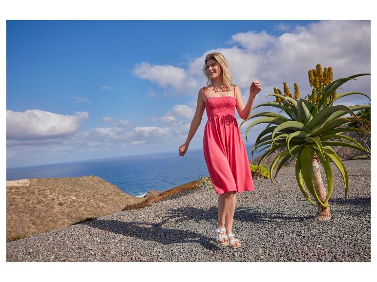 Vrouw in een roze zomerjurk en sandalen, buiten staand aan zee met een aloëplant.
