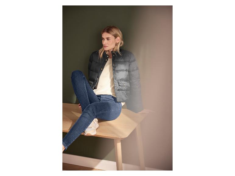 Vrouw in zwarte gewatteerde jas, crèmekleurig T-shirt en blauwe jeans, zittend op een houten bankje.