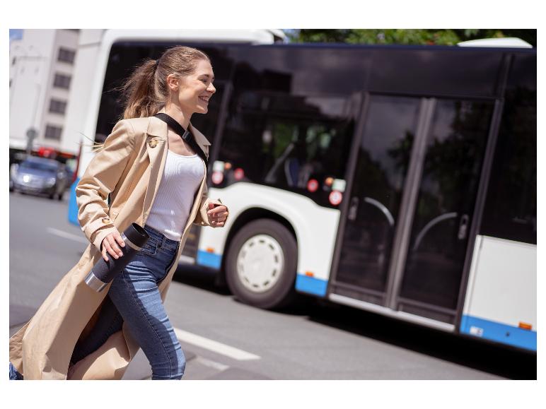 Lachende vrouw in trenchcoat en jeans, met reismok, rent langs een bus.