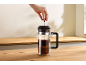 Een hand drukt de zuiger van een koffiepers met koffie naar beneden op een houten aanrecht.
