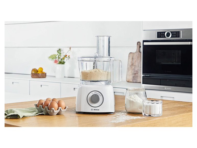 Een witte Bosch keukenmachine met een kom vol deeg, eieren en bloem op een houten aanrecht.
