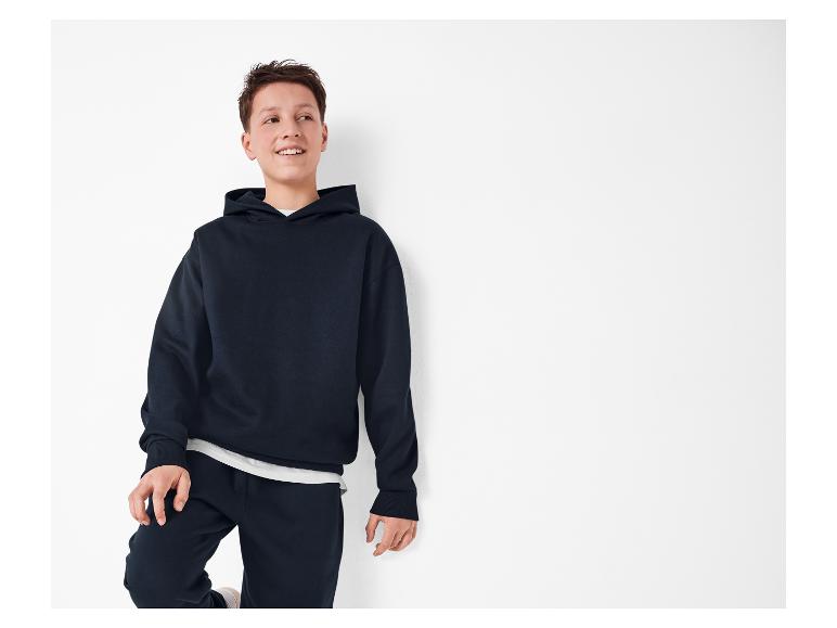 Een jongen in een donkerblauwe hoodie en joggingbroek lacht terwijl hij tegen een witte muur leunt.