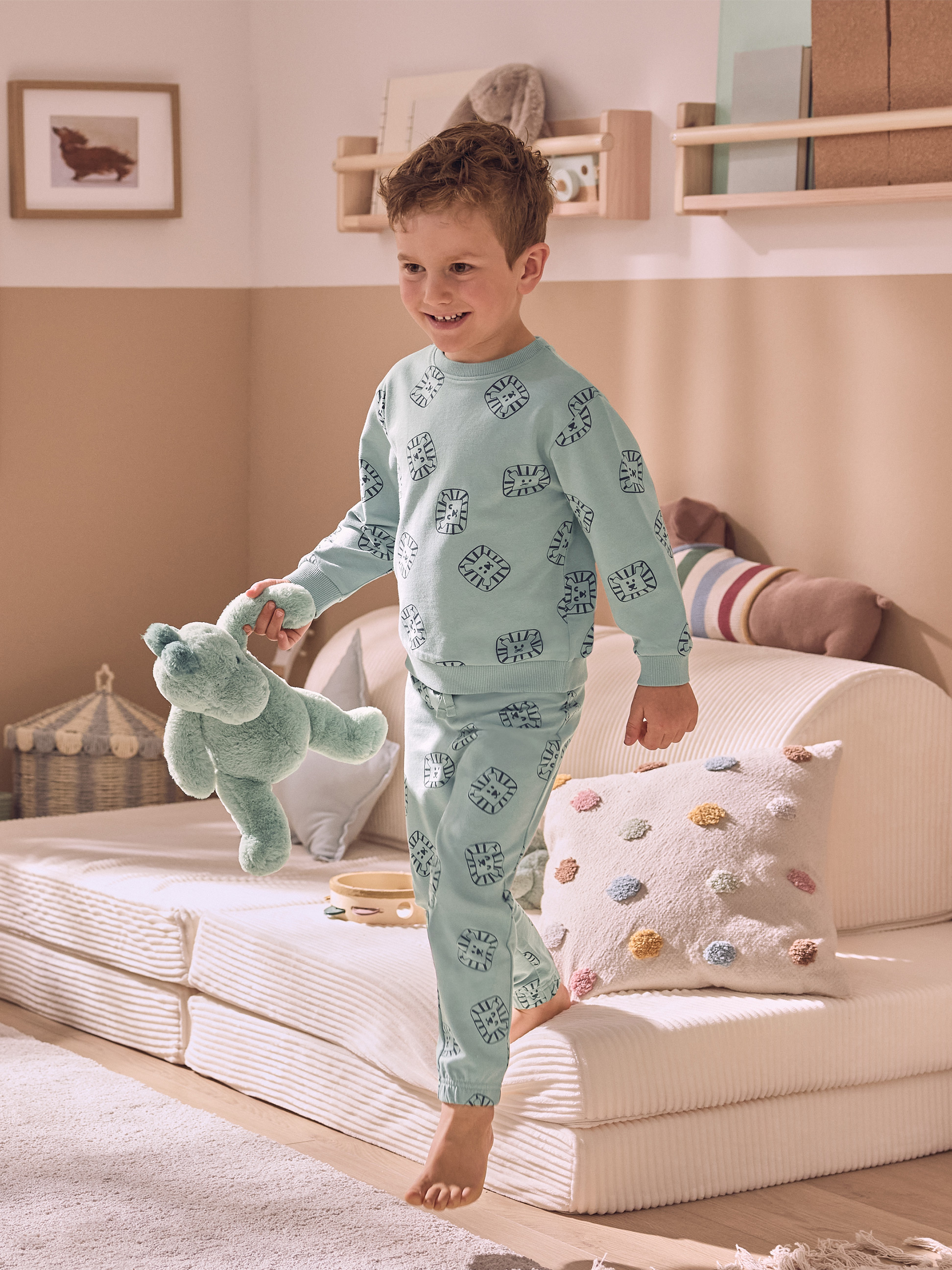 Een lachend kind in lichtblauwe pyjama met leeuwenmotieven, dat een groen knuffeldier vasthoudt.