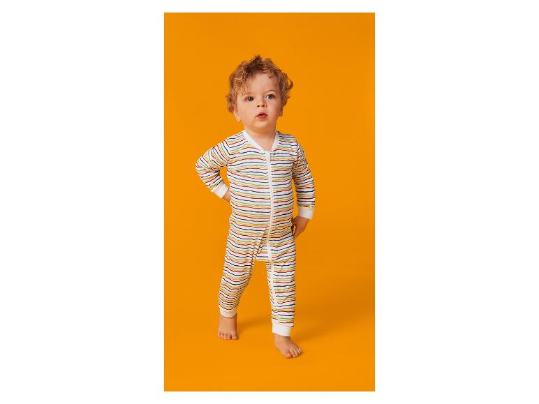 Krullendharige baby in gestreepte romper staat op een oranje achtergrond.