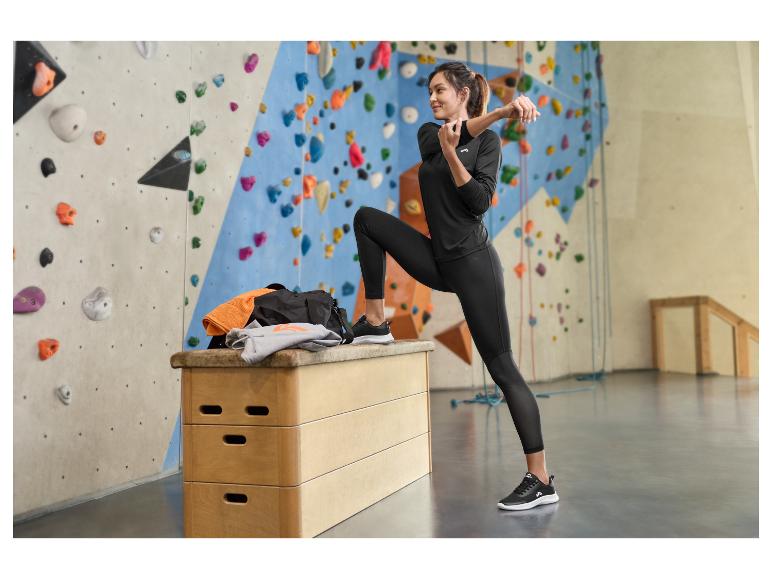 Vrouw in zwarte sportkleding rekt zich uit naast een klimmuur met sporttas en hoodie.