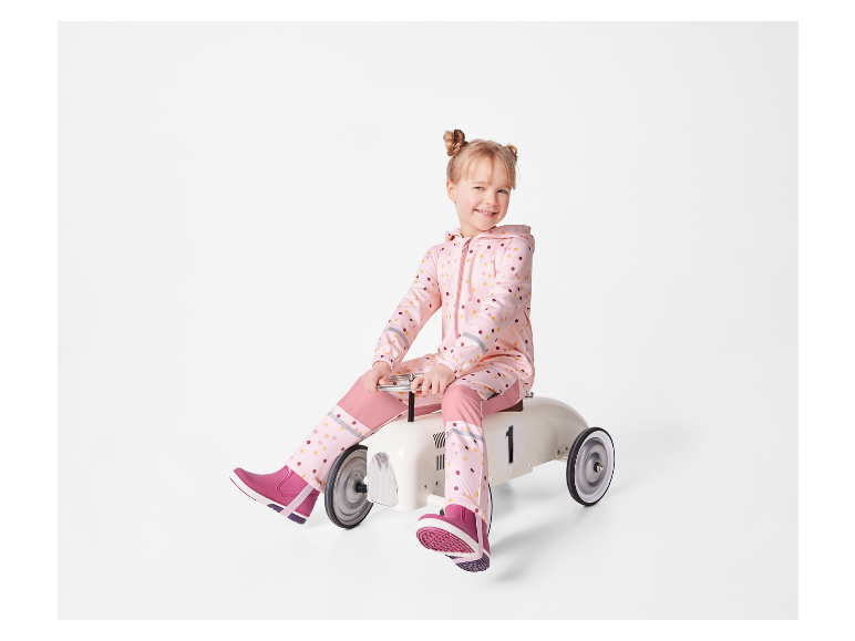 Een lachend meisje in een roze jumpsuit met stippen, rijdt op een wit speelgoedautootje.