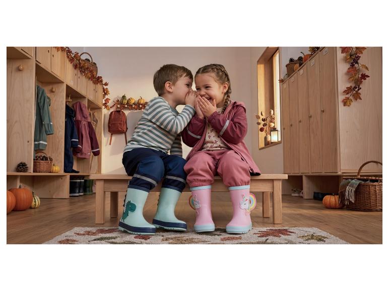Twee kinderen in regenpakken en regenlaarzen fluisteren geheimen in een herfstgarderobe.