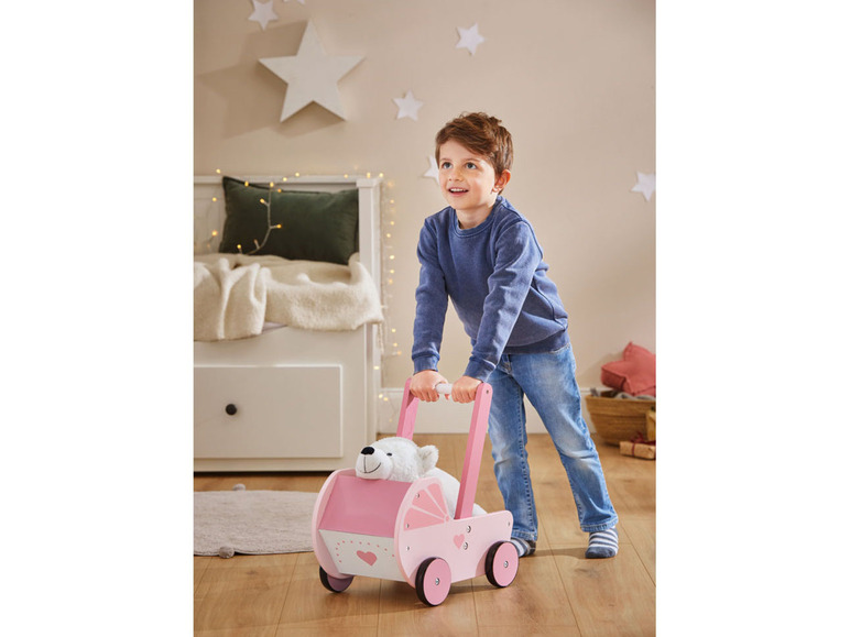 Een jongetje duwt een roze houten loopwagen met een knuffelbeer erin.