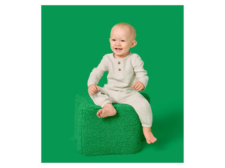 Lachende baby in een beige mousseline set, zittend op een groene poef.