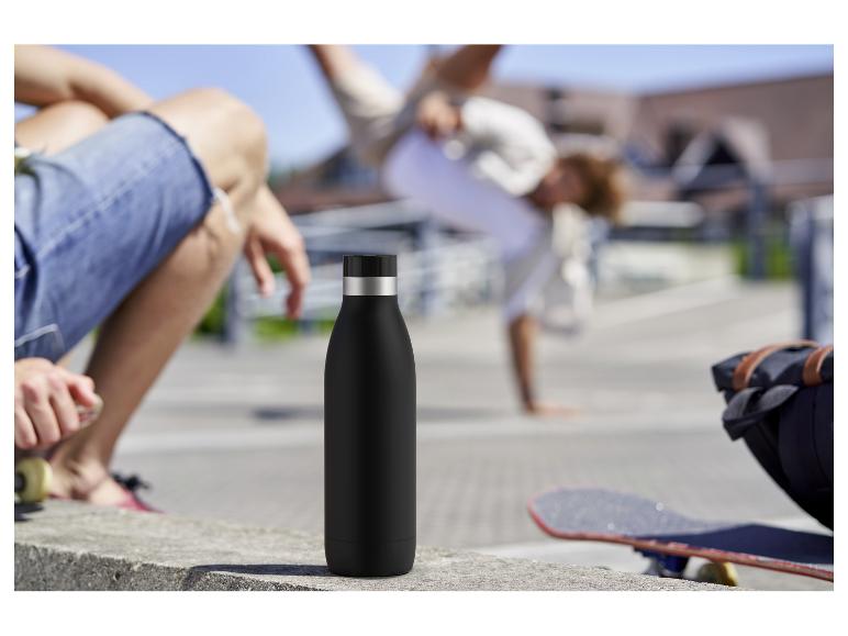 Zwarte geïsoleerde fles op een betonnen rand met mensen en een skateboard op de achtergrond.