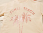 Een beige t-shirt met de tekst 'Sunset Beach' en palmbomen.