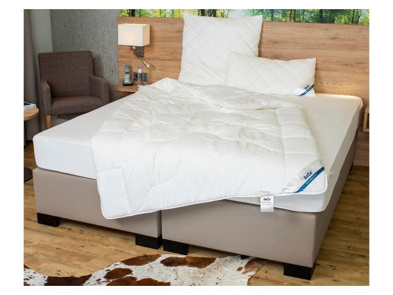 Bed met wit matras, BeCo gewatteerde dekbedden en kussens, met een fauteuil en nachtkastje.