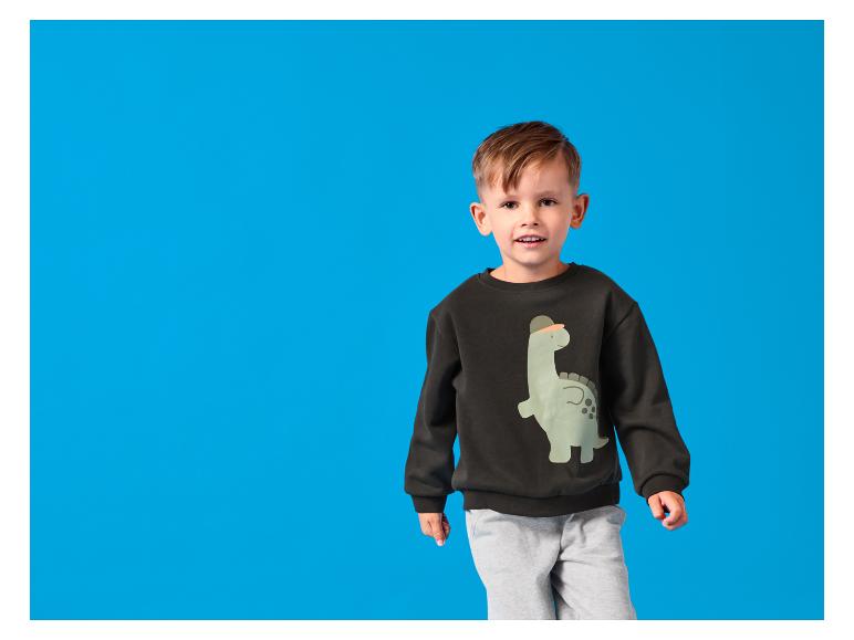 Blije jongen in zwarte sweater met dinoprint en grijze joggingbroek.