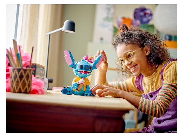 Een lachend meisje speelt met een grote Stitch-figuur gemaakt van LEGO-stenen op een tafel.