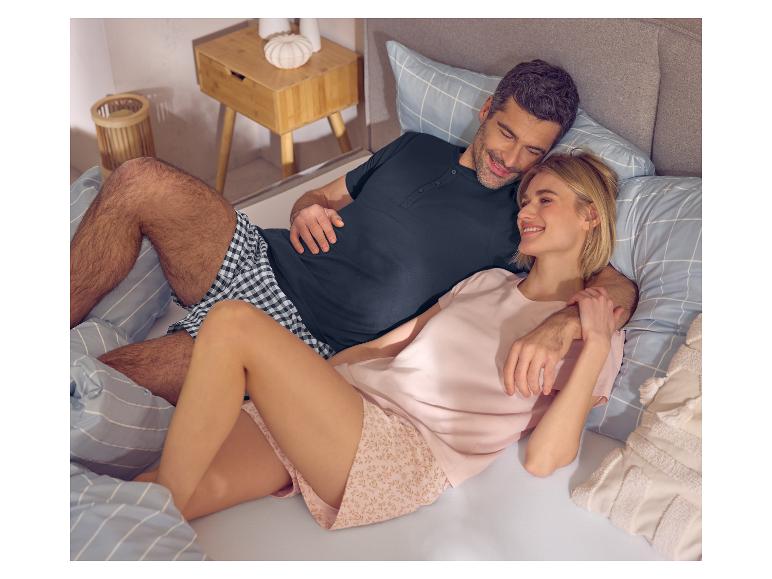 Lachend stel in pyjama in bed, man in donkere top en geruite shorts, vrouw in roze top en bloemen shorts.