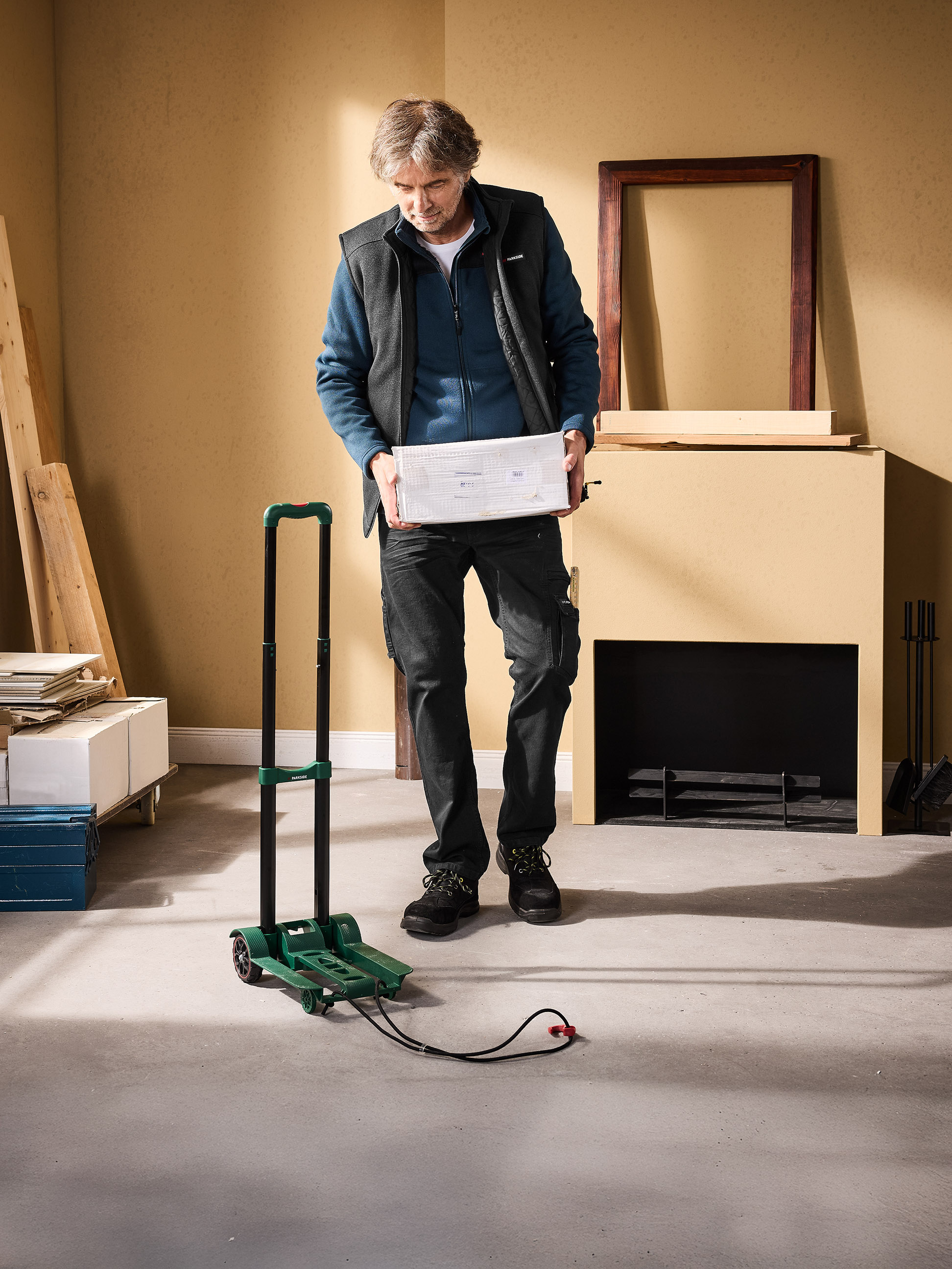 Man met een witte doos, naast een groene trolley en een open haard.