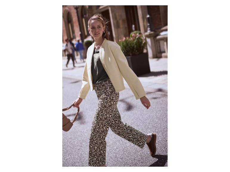 Vrouw in lichte jas, groen T-shirt en luipaardprint broek loopt op straat.